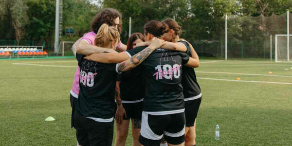 Devojke na sportskom terenu u crnim dresovima sa belo-roze natpisom zagrljene sa čovekom koji nosi roze majicu.