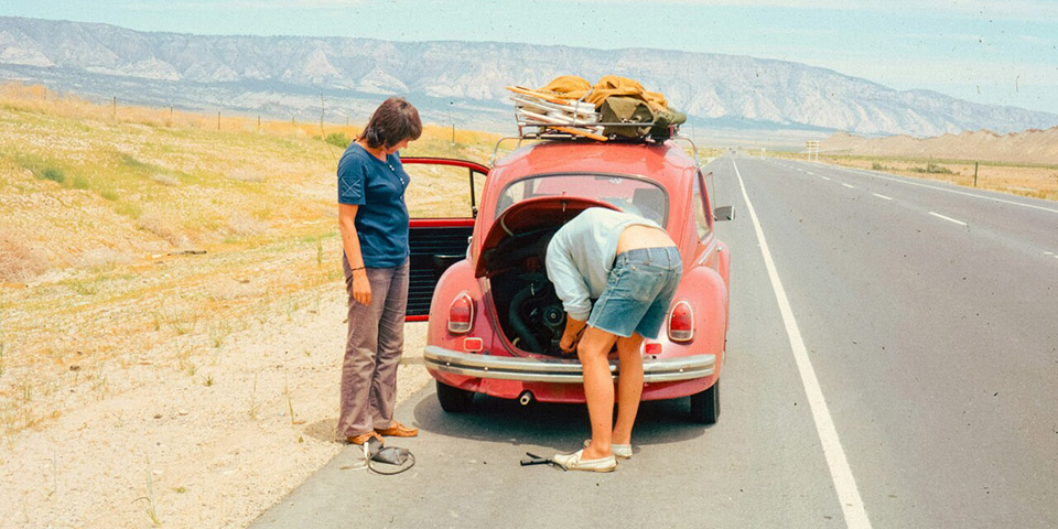 Dvoje ljudi pored starog crvenog automobila na putu dok jedan proverava otvoren prtljažnik