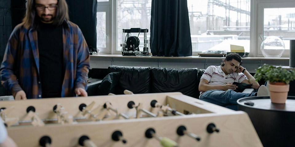 Muškarac leži na crnom kauču i gleda telefon dok drugi stoji pored stonog fudbala