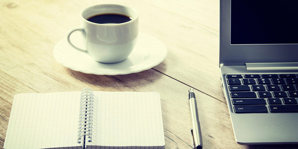 Otvorena sveska na kvadratić, hemijska olovka, šolja kafe i laptop na drvenom stolu
