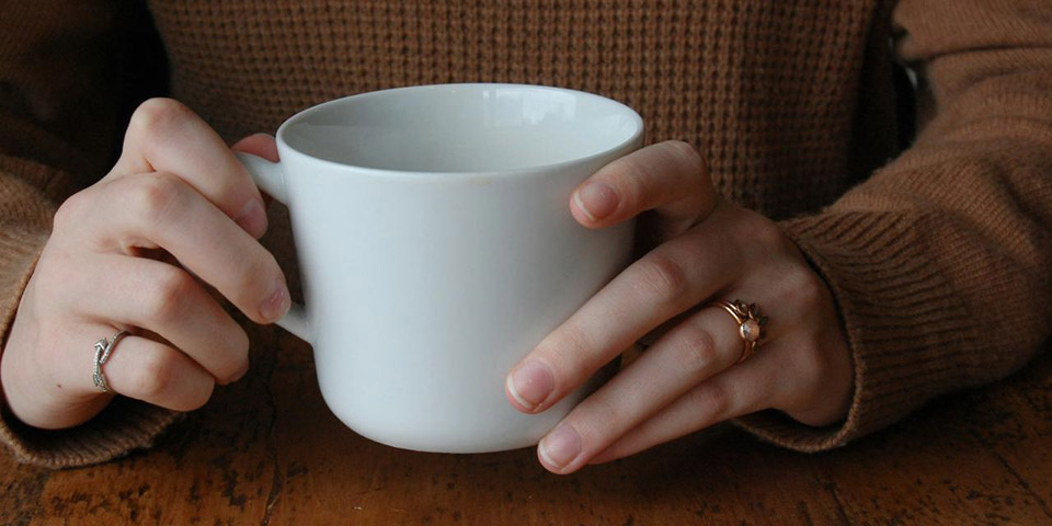 Ruke u smeđem džemperu drže veliku belu šolju na drvenom stolu
